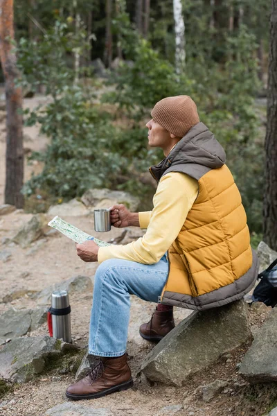 Hiker in warm vest and beanie looking away while sitting with mug and map in forest — Stock Photo
