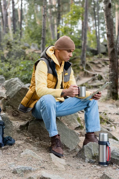 Touriste regardant la carte tout en étant assis avec une tasse en métal dans la forêt — Photo de stock