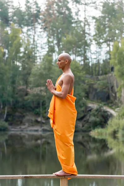 Budista descalzo de pie sobre una valla de madera en el bosque mientras medita con las manos orantes - foto de stock