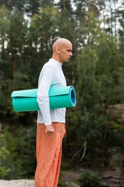 Side view of buddhist in white sweatshirt standing with yoga mat in forest — Stock Photo