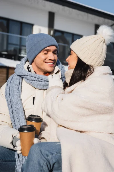 Allegro uomo e donna in cappelli a maglia che si guardano mentre tengono in mano bicchieri di carta — Foto stock