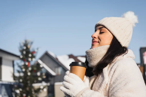 Mujer alegre con los ojos cerrados y sombrero de invierno sosteniendo taza de papel afuera - foto de stock