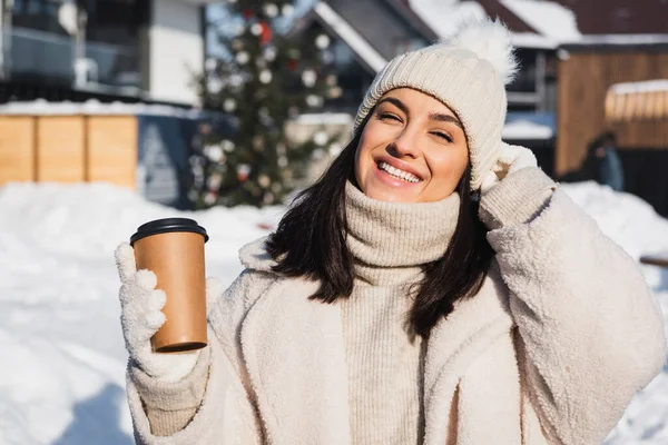 Mujer joven feliz en sombrero de punto sosteniendo taza de papel afuera - foto de stock