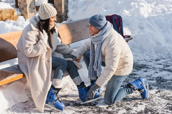 Happy man wearing ice skates on cheerful woman in winter hat — Stock Photo