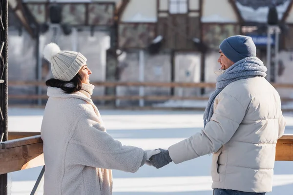 Feliz jovem casal em chapéus de inverno olhando um para o outro, mantendo as mãos fora — Fotografia de Stock