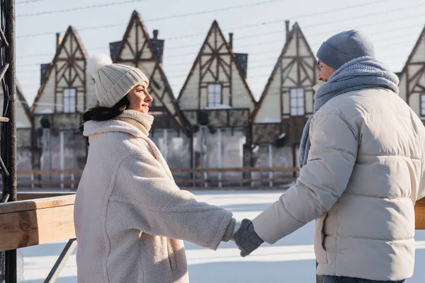 Веселая молодая пара в зимних шляпах смотрит друг на друга, держа за руки снаружи — стоковое фото