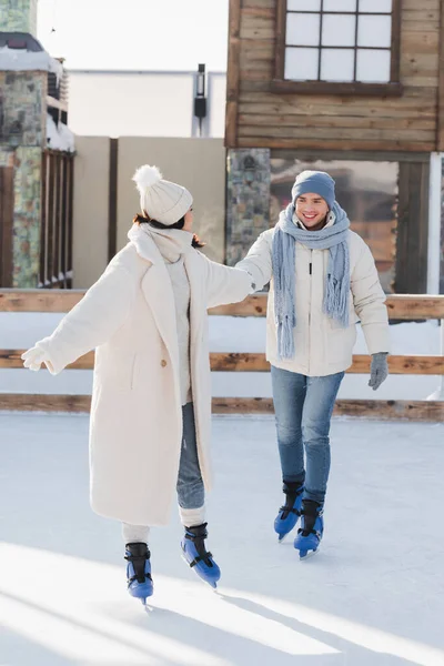 In voller Länge glücklicher junger Mann mit Wintermütze und Schlittschuhen hält Händchen mit Freundin auf Eisbahn — Stockfoto