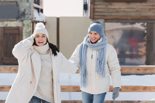 Happy young man in winter hat holding hands with girlfriend on ice rink — Stock Photo