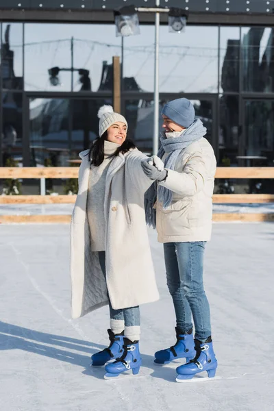 Full length of happy young man in winter hat and smiling girlfriend with outstretched hand skating on ice rink — Stock Photo