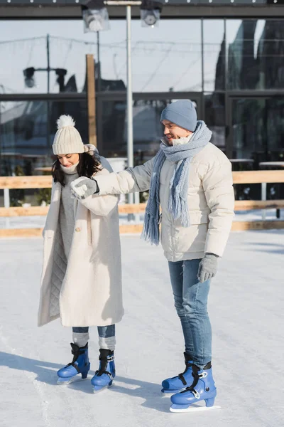 Comprimento total do jovem feliz em chapéu de inverno e mulher patinando na pista de gelo — Fotografia de Stock