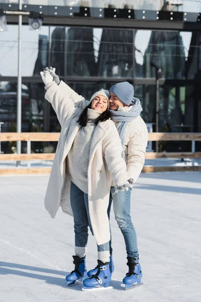 Pleine longueur de heureux jeune homme et femme en chapeaux d'hiver patiner sur la patinoire — Photo de stock