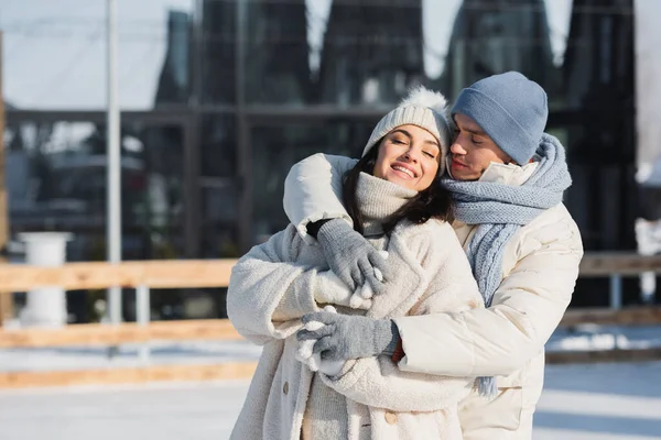 Happy young man and woman in winter hats hugging on ice rink — Stock Photo