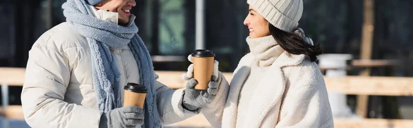 Fröhlicher junger Mann schenkt Pappbecher an fröhliche Freundin mit Wintermütze, Banner — Stockfoto