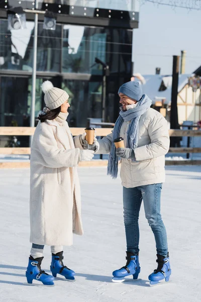 Pleine longueur de jeune homme heureux donnant tasse en papier à petite amie gaie en chapeau d'hiver sur la patinoire — Photo de stock