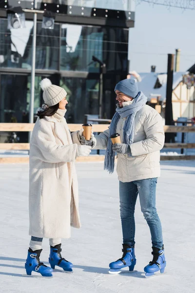 Full length of happy young man giving paper cup to cheerful girlfriend in winter hat on ice rink — Stock Photo