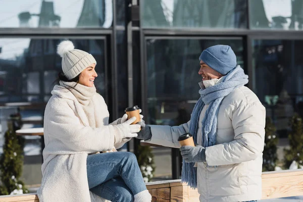 Fröhlicher junger Mann schenkt Pappbecher an fröhliche Freundin mit Wintermütze — Stockfoto