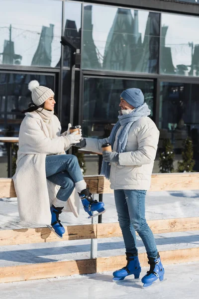 Pleine longueur de jeune homme heureux donnant tasse en papier à petite amie en chapeau d'hiver près de la patinoire — Photo de stock