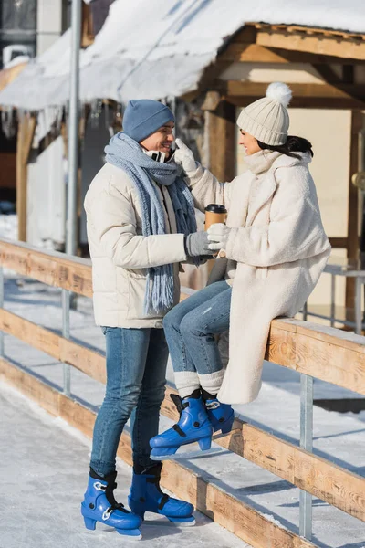 Full length of happy young man holding paper cup near cheerful girlfriend in winter hat and ice skates near ice rink — Stock Photo