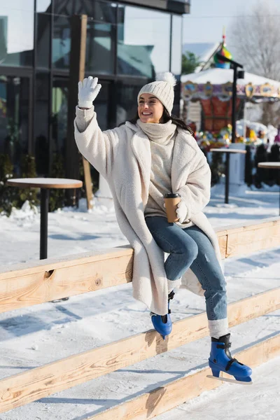 Piena lunghezza di felice giovane donna in inverno cappello e pattini da ghiaccio tenendo tazza di carta e agitando la mano vicino alla pista di pattinaggio — Foto stock