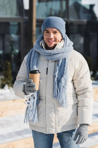 Felice giovane uomo in inverno cappello e sciarpa tenendo tazza di carta al di fuori — Foto stock