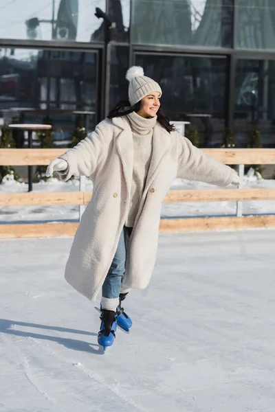 Pleine longueur de jeune femme heureuse en chapeau d'hiver et manteau patinant sur la patinoire — Photo de stock