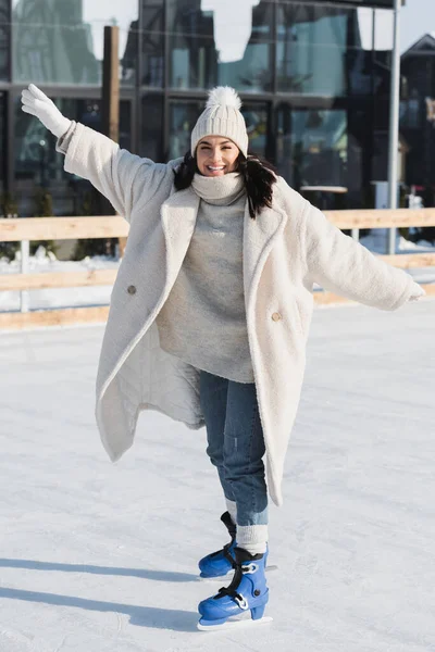 Longitud completa de la joven complacida en sombrero de invierno y capa de patinaje en pista de hielo - foto de stock