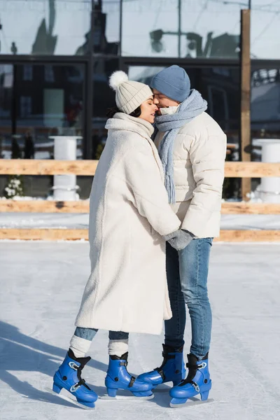 Full length of young couple in winter hats holding hands while kissing on ice rink — Stock Photo