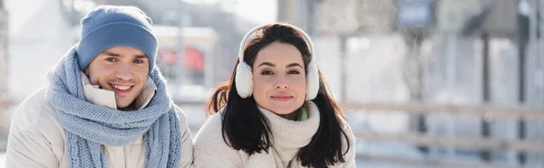 Feliz jovem mulher no ouvido muffs e homem alegre no chapéu de inverno olhando para a câmera fora, banner — Fotografia de Stock