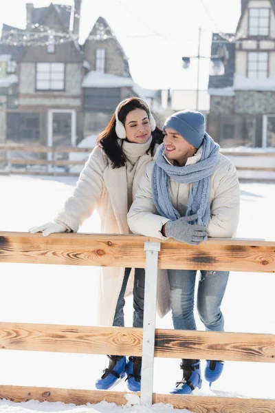 In voller Länge eine glückliche junge Frau mit Ohrenschützern, die einen fröhlichen Mann mit Wintermütze ansieht, der sich an eine Holzkante auf der Eisbahn lehnt — Stockfoto