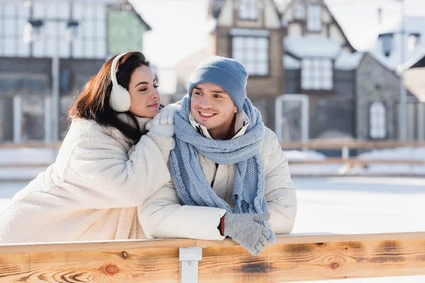 Young woman in ear muffs and cheerful man in winter hat leaning on wooden border on ice rink — Stock Photo