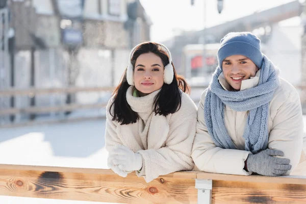 Fröhliche junge Frau mit Ohrenschützer und fröhlicher Mann mit Wintermütze, angelehnt an Holzrahmen auf der Eisbahn — Stockfoto