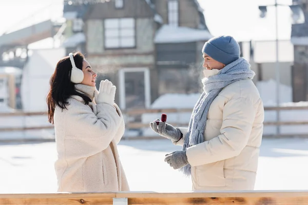 Vue latérale de l'homme en chapeau d'hiver tenant boîte avec anneau de mariage près de la jeune femme avec les yeux fermés — Photo de stock