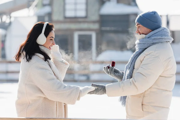 Вид сбоку мужчины в зимней шляпе, держащего коробку с обручальным кольцом рядом со счастливой молодой женщиной в наушниках — стоковое фото