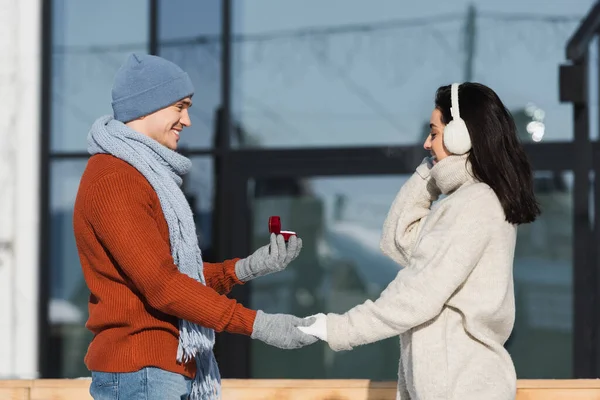 Вид сбоку улыбающегося мужчины в зимней шляпе, держащего коробку с обручальным кольцом рядом со счастливой молодой женщиной в наушниках — стоковое фото