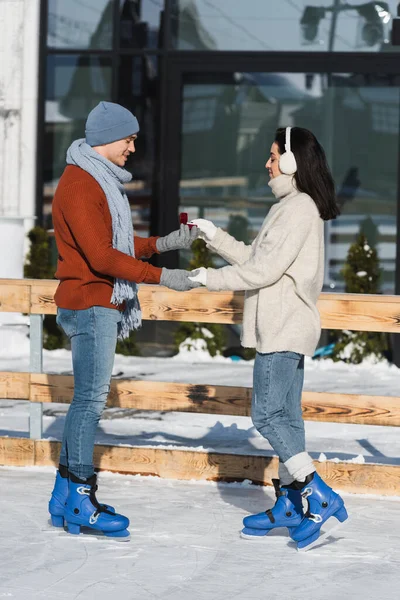 Vista lateral del hombre sonriente en caja de celebración de sombrero de invierno con anillo de boda cerca de mujer joven en pista de hielo — Stock Photo