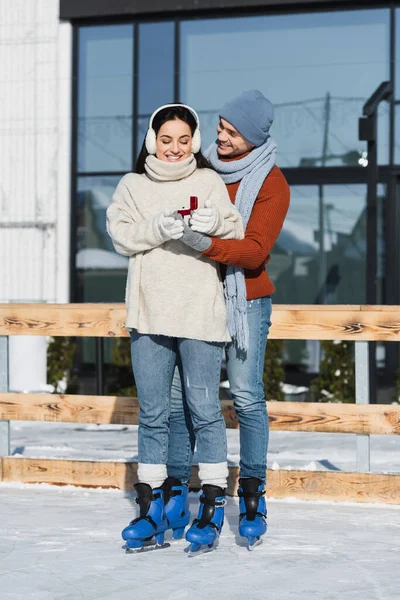 Volle Länge des lächelnden Mannes mit Wintermütze, der Schachtel mit Ring hält, während er der glücklichen Frau im Ohrenschutz auf der Eisbahn einen Heiratsantrag macht — Stockfoto