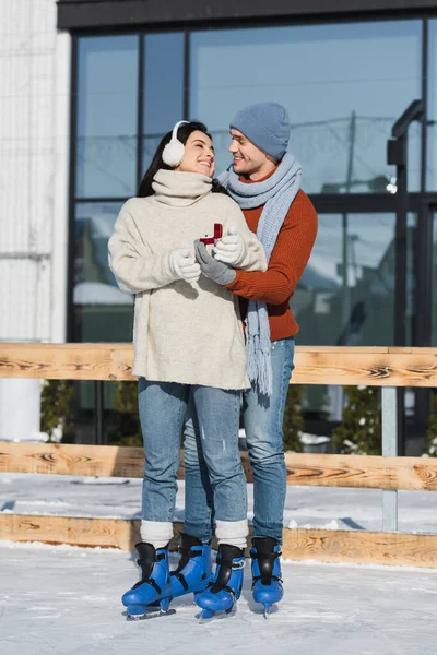 Full length of cheerful man in winter hat holding box with ring while making proposal to young woman in ear muffs on ice rink — Stock Photo