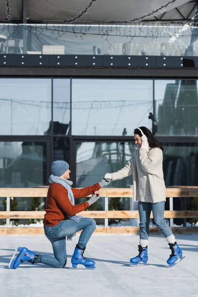Pleine longueur de l'homme agenouillé et tenant la boîte avec anneau tout en faisant une proposition à la femme heureuse dans des cache-oreilles sur la patinoire — Photo de stock