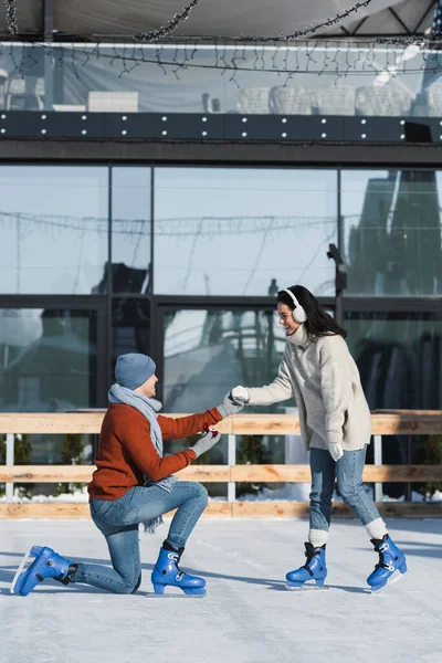 Full length of young man holding box with ring and kneeling while making proposal to happy woman in ear muffs on ice rink — Stock Photo