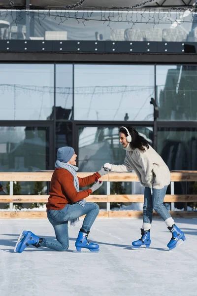 Volle Länge des Mannes hält Box mit Ehering und kniet, während er der erstaunten Frau im Ohrenschützer auf der Eisbahn einen Heiratsantrag macht — Stockfoto