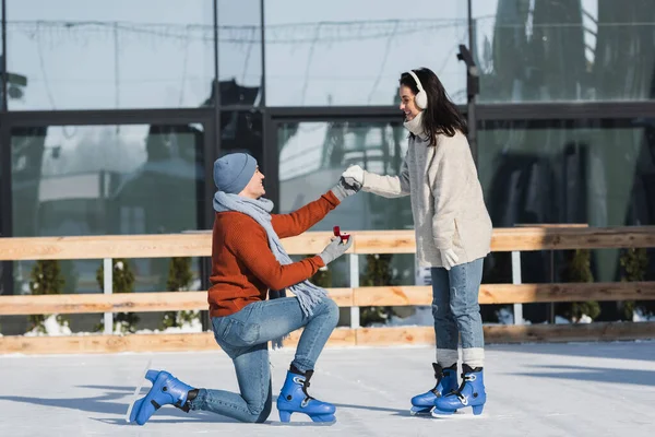 Homme tenant boîte avec anneau de mariage et à genoux tout en faisant une proposition à la femme heureuse dans des cache-oreilles sur la patinoire — Photo de stock