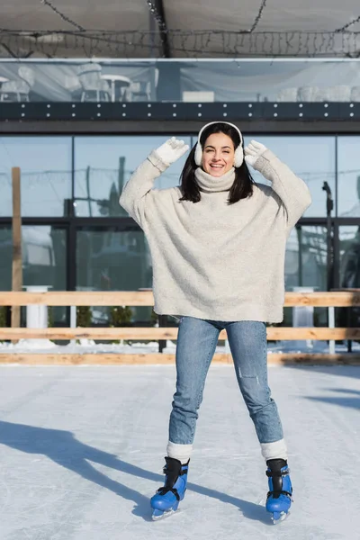 Pleine longueur de joyeuse jeune femme en pull et cache-oreilles patiner sur la patinoire — Photo de stock