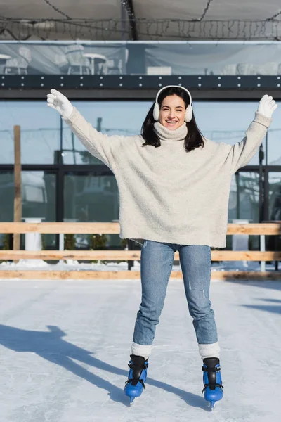 Piena lunghezza di felice giovane donna in maglione e cuffie pattinaggio su pista di pattinaggio su ghiaccio — Foto stock