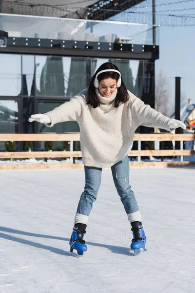 Full length of young woman in sweater and ear muffs smiling while skating on ice rink — Stock Photo