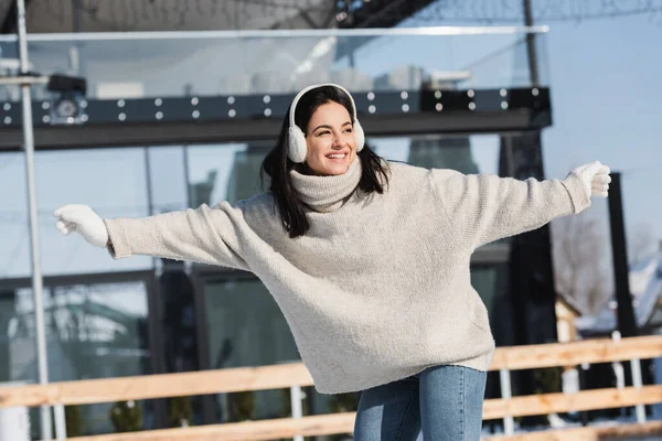 Jeune femme en pull et cache-oreilles souriant tout en patinant sur la patinoire — Photo de stock