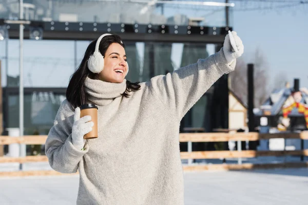 Glückliche Frau in Pullover und Ohrenschützer hält Pappbecher in der Hand und macht Selfie auf der Eisbahn — Stockfoto