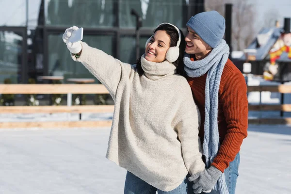 Mujer feliz en suéter y orejeras tomando selfie con novio en pista de hielo - foto de stock