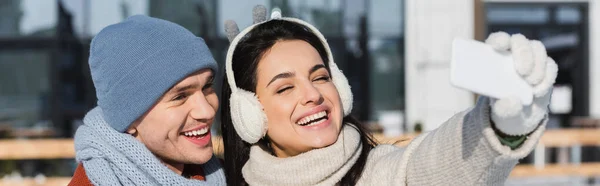 Mujer feliz en suéter y orejeras tomando selfie con novio en invierno, pancarta - foto de stock