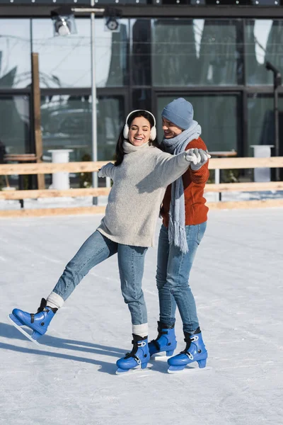 Longitud completa de la mujer feliz en orejeras cogidas de la mano con el novio alegre mientras patina en pista de hielo - foto de stock