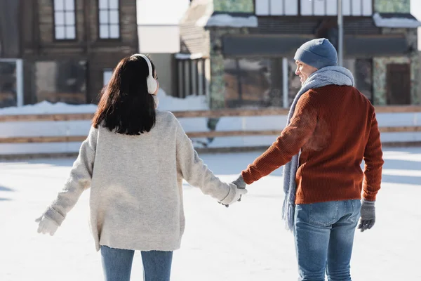 Brunette young woman in ear muffs holding hands with cheerful boyfriend on ice rink — Stock Photo